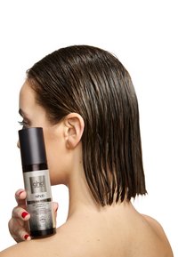 GHD Rehab tunna hår förtjockningsspray i en klar flaska med svart lock, hålls nära blött, rakt medelbrunt hår. Slät textur, minimalistisk design.