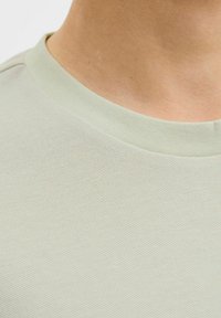 Tricou de culoare verde deschis, cu un model texturat și guler rotund, fabricat din material respirabil. Prim-planul arată gulerul și un model țesut subtil.