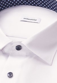 Close-up van de kraag van een witte dress shirt met donkere knopen en een label met de tekst "seidensticker," dat blauwe stof met lichte stippen aan de binnenkant van de kraag toont.