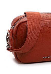 Kleine crossbodytas van roestkleurig, gestructureerd leer met brede stoffen band, zilveren rits en metalen merklogo "SURI FREY."