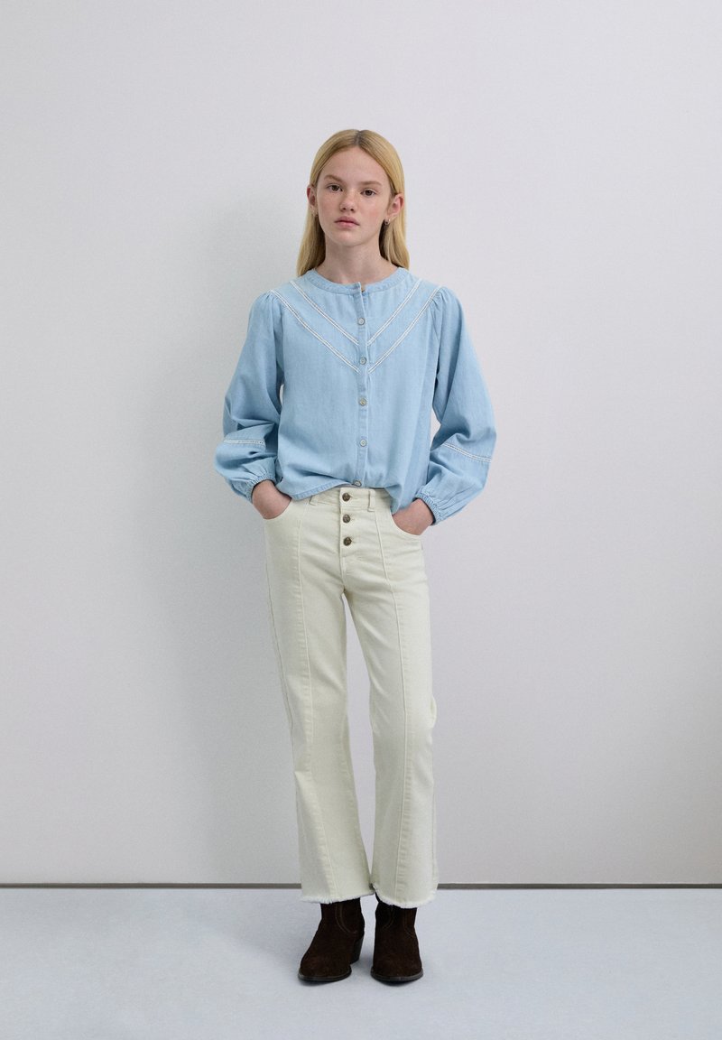 Blusa azul claro abotonada con mangas abullonadas y detalles bordados en blanco, combinada con jeans de pierna ancha color crema de talle alto y botas marrones hasta el tobillo.