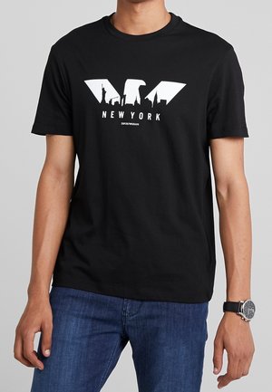 Man wearing black T-shirt with white New York skyline graphic and text, paired with blue jeans and wristwatch with black strap.