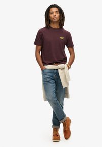 Bordowa koszulka z małym żółtym logo, jasnoniebieskie dżinsy i brązowe buty. Sweter związany w talii dodaje faktury i kontrastu.