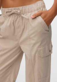 Beige cargobroek met een elastische tailleband, zijzakken en een met een knoop gesloten flapzak op het been. Gemaakt van lichtgewicht, soepele stof.