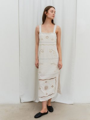 Donna che indossa un abito midi bianco senza maniche ricamato con una spacco, in piedi davanti a una tenda bianca semplice, guardando di lato.