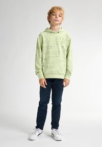 Sudadera verde claro con patrones geométricos negros, de mangas largas y con un bolsillo frontal. Combinada con vaqueros azul oscuro y zapatillas blancas.