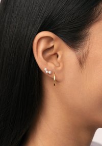 Pendiente de aro dorado con tres pequeñas perlas blancas que realzan la oreja. Textura de metal suave con un acabado brillante. Diseño simple y moderno.
