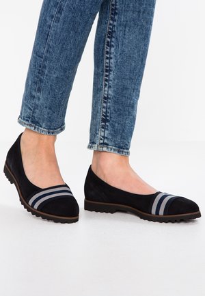 Fötter i svarta slip-on skor med grå ränder på tårna, klädda i blå denimjeans, står på ett vitt golv.
