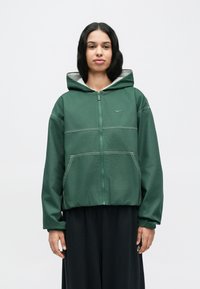 Sweat à capuche zippé vert avec une capuche doublée de gris, présentant des accents de coutures blanches et deux poches avant. Tissu texturé, coupe décontractée.