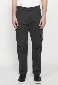 Zwarte cargobroek met meerdere zijzakken, een aansluitend ontwerp en een gladde textuur, gecombineerd met zwarte sneakers op een plain achtergrond.