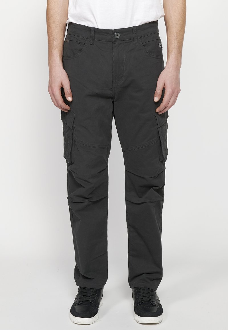 Zwarte cargobroek met meerdere zijzakken, een aansluitend ontwerp en een gladde textuur, gecombineerd met zwarte sneakers op een plain achtergrond.