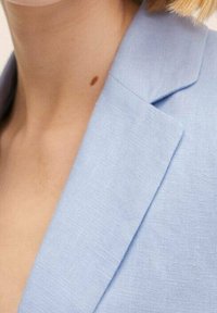 Blazer di lino azzurro chiaro con colletto a revers e spalle strutturate. La texture è leggermente ruvida, con dettagli di cucitura visibili.