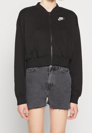 Persoon draagt een zwart Nike jack met rits en lange mouwen en donkergrijze denim shorts met hoge taille en rafelige zoom.