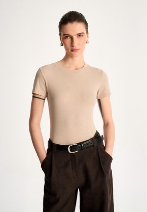 Młoda osoba z zaczesanymi do tyłu włosami, ubrana w beżową bluzkę z krótkim rękawem, ciemnobrązowe spodnie z wysokim stanem oraz czarny pasek i złote kolczyki koła.