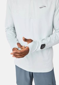 Šviesiai mėlynas ilgomis rankovėmis sportinis marškinėlis su pusiau užtraukiamu apykakle ir nykščių angomis, derinamas su pilkais šortais ir juodu laikrodžiu ant riešo.