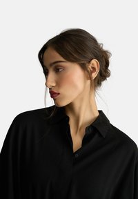 Czarna koszula z guzikami i kołnierzykiem, wykonana z gładkiego materiału. Posiada luźny krój i długie rękawy. Włosy modelki ułożone w kok.