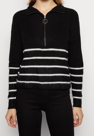 Schwarzer Strickpullover mit weißen horizontalen Streifen am Körper und an den Ärmeln, mit einem halbhohen Reißverschluss mit rundem Zipper, kombiniert mit schwarzen Hosen.