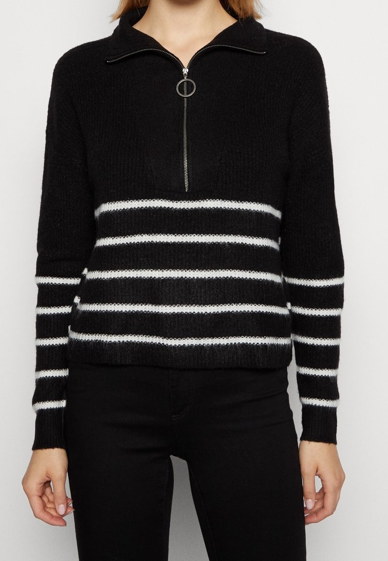 Pull en maille noire avec des rayures horizontales blanches sur le corps et les manches, doté d'une fermeture à glissière à moitié fermée avec un tire-zip circulaire, porté avec un pantalon noir.