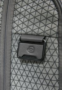 Zaino grigio con trama testurizzata e motivo geometrico a triangoli, con una cinghia in pelle nera come accento e una targhetta con il logo. Cerniera visibile.