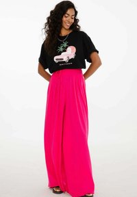 Žena s dlouhými kudrnatými vlasy, oblečená v černém tričku s potiskem a jasně růžových širokých kalhotách, stojící s rukama v bok a usmívající se dolů.