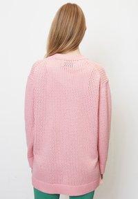 Světle růžový pletený svetr s volným střihem, který má strukturovaný, otevřený vzor a vroubkovaný lem. Dlouhé rukávy a design s kulatým výstřihem.
