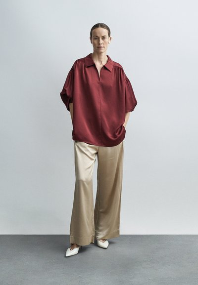 Kvinde iført en løs burgunderfarvet bluse, beige vidde bukser i satin, hvide spidse sko og et beige høreapparat, stående mod en lysegrå baggrund.