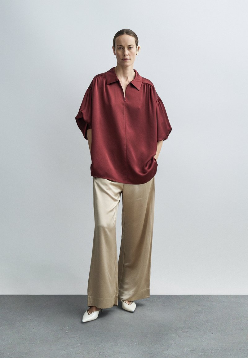 Woman wearing a loose burgundy blouse, beige wide-leg satin pants, white pointed shoes, and a beige hearing aid standing against a light gray background.