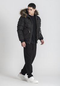 Giacca nera imbottita con cappuccio in pelliccia sintetica, tasche con zip e una texture liscia, abbinata a pantaloni neri e sneakers bianche.