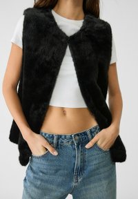 Kobieta w czarnej kamizelce z sztucznego futra nosi białą bluzkę crop top i ma ręce w kieszeniach niebieskich dżinsów, na tle jednolitego tła.