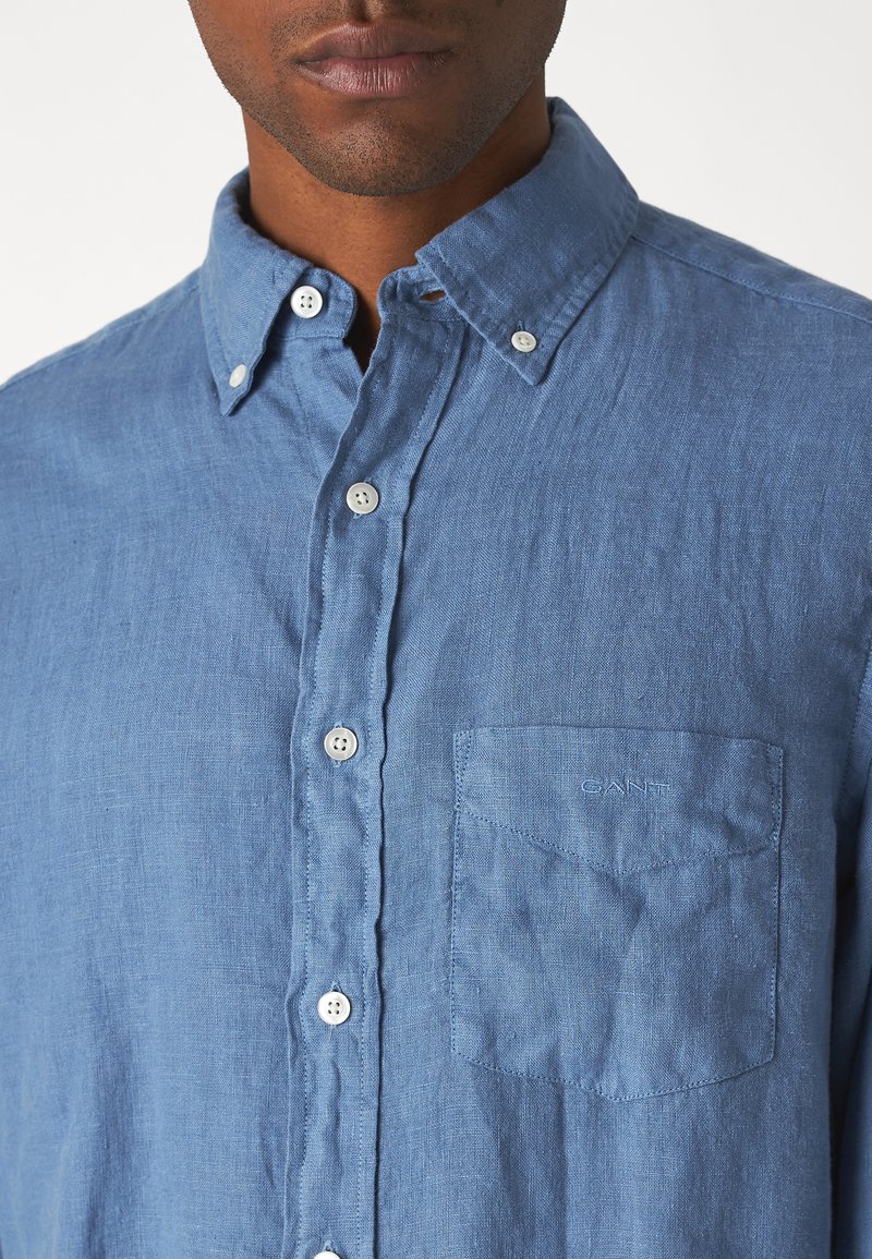 Helles blaues Leinenhemd mit Button-Down-Kragen, weißen Knöpfen und einer Brusttasche mit subtiler Logo-Stickerei. Strukturierter Stoff.