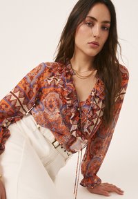 Vrouw met lang donker haar, gekleed in een bloemenpatroon oranje en blauwe blouse met ruches en een witte broek met hoge taille, poserend tegen een effen achtergrond.