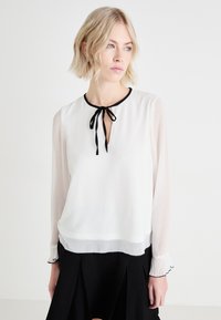 Blouse blanche avec des manches longues transparentes et un col noir. Présente un décolleté en forme de clef à l'avant et un détail de nœud au niveau du col. Poignets texturés avec des accents noirs.