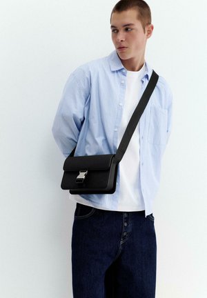 Young man in light blue shirt and white tee wearing black crossbody bag, looking sideways against a plain light background.