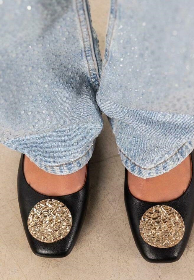 Zapatos negros de punta cuadrada con grandes acentos circulares dorados, combinados con jeans de denim azul claro adornados.