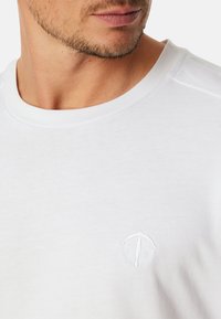 Camiseta de algodón blanca con un cuello acanalado y mangas cortas; presenta un pequeño logo bordado en el pecho. Textura suave.