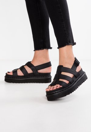 Pieds portant des sandales à plateforme noires épaisses avec plusieurs brides et boucles, assorties à un pantalon noir à ourlet effiloché.