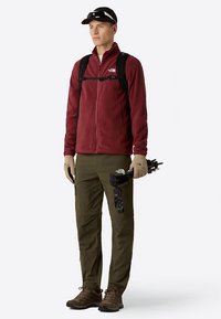 Giacca rossa in pile con zip, pantaloni cargo verdi oliva, stivali da trekking marroni, cappellino nero e zaino; tiene in mano bastoncini da trekking neri.