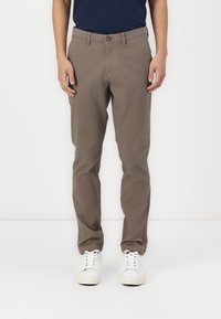 Braune Baumwollhosen mit Flachfront, Gürtelschlaufen und Knopfverschluss. Kombiniert mit weißen Sneakers, vor einem schlichten Hintergrund stehend.
