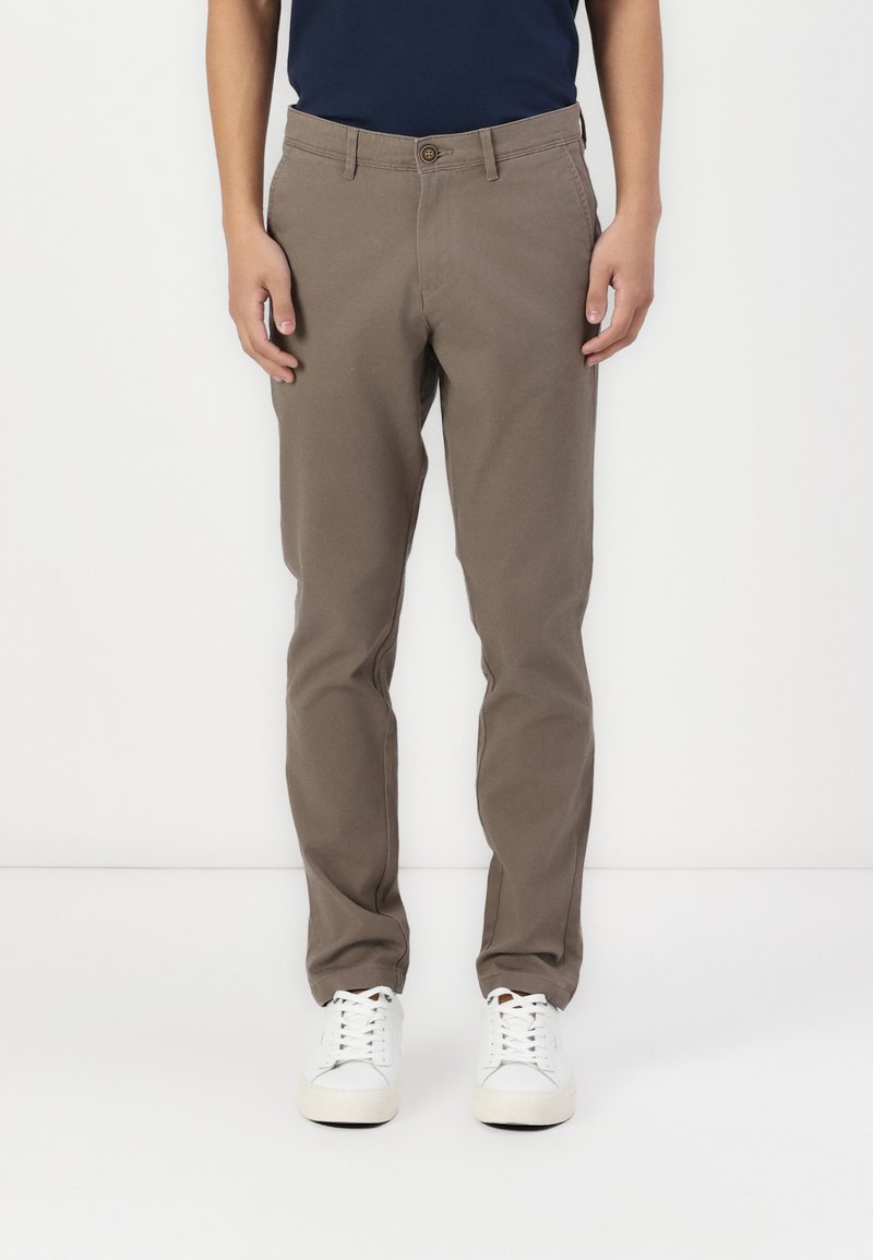 Braune Baumwollhosen mit Flachfront, Gürtelschlaufen und Knopfverschluss. Kombiniert mit weißen Sneakers, vor einem schlichten Hintergrund stehend.