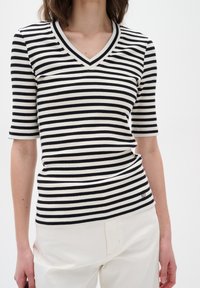 Frau trägt ein schwarz-weiß gestreiftes V-Ausschnitt-T-Shirt mit kurzen Ärmeln, kombiniert mit hochtaillierten weißen Hosen, sichtbar von Hals bis Hüfte.