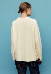 Cremefarbener, übergroßer Pullover mit weicher Textur, mit tiefer angesetzten Schultern und einem gerippten Saum, darüber getragen ein dunkles Oberteil.