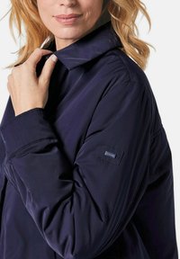 Navyblaue, leichte Jacke mit Kragen, die eine glatte Textur aufweist und ein Logoschild auf dem Ärmel hat. Verstellbare Bündchen und eine lockere Passform.