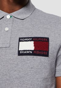 Szary podkoszulek z materiałową łatą z napisem "TOMMY HILFIGER" i "SHAWN MENDES", z czerwonymi i białymi akcentami oraz zapięciem na guziki.