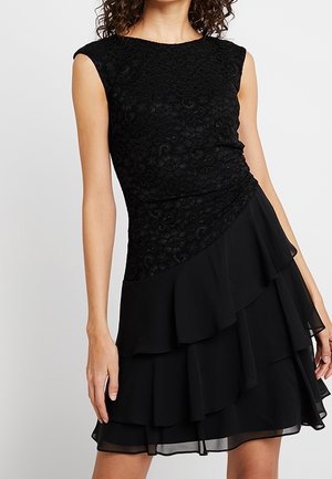 Kvinde iført ærmeløs sort kjole med tekstureret blondeoverdel og lagdelt chiffon nederdel mod en ensfarvet lys baggrund.
