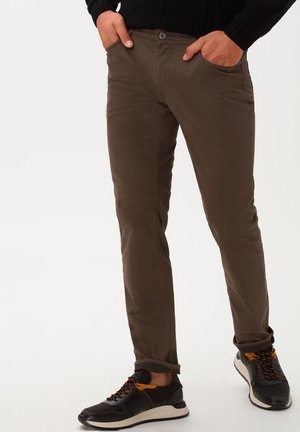 Pantaloni marroni slim fit indossati con sneakers nere e arancioni, mani appoggiate nelle tasche davanti su uno sfondo chiaro e uniforme.