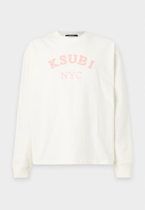 Man die bruine zonnebril draagt, wit shirt met lange mouwen met tekst "K SUBI NYC", lichtblauwe versleten spijkerbroek en zwarte enkellaarzen, staand tegen een witte achtergrond.