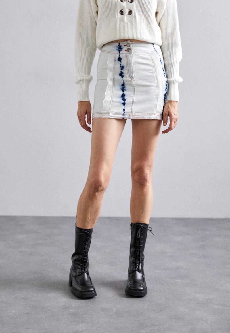 Jupe en denim mini bleue claire avec un motif tie-dye et des coutures contrastées. Portée avec des bottines noires à lacets.