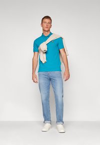 Türkiissinine polo särk lühikeste varrukatega, väikese tikitud logoga, koos helesiniste teksase ja valgeid tennisetossudega. Beez kampsun õlgadel.