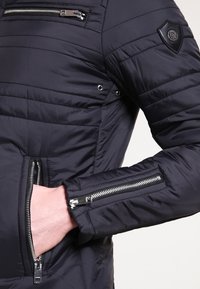 Schwarze Steppjacke mit einem gesteppten Design, Reißverschluss-Brust- und Handgelenktaschen sowie einem strukturierten Schulterpatch für zusätzlichen Details.