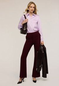 Kvinde med blondt hår iført en lilla satinbluse, mørkerøde bukser, sorte hæle, der holder en sort læderjakke og skuldertaske.